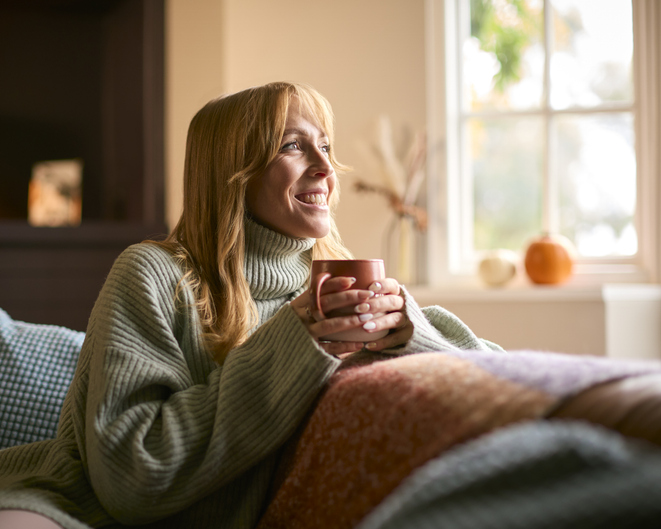 Smiling Woman At Home Wearing Winter Jumper With Warming Hot Drink Of Tea Or Coffee In Cup Or Mug