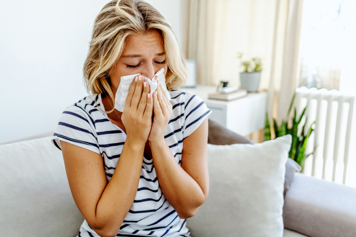 Shot of a young woman feeling ill and blowing her nose with a tissue at home. Photo of sneezing woman in paper tissue. Picture showing woman sneezing on tissue on couch in the living-room.