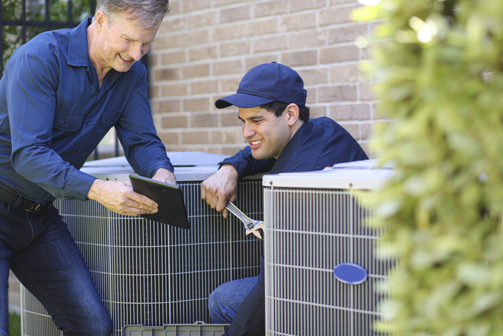blue collar air conditioner repairmen working at residential home. They prepare to begin work by gathering appropriate tools and referring to digital tablet.